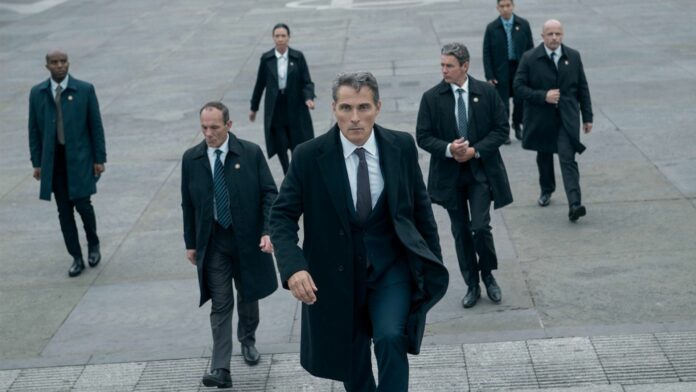 Rufus Sewell walks upstairs wearing a suit surrounded by other people wearing dark suits in a still from Netflix&#039;s The Diplomat season 3