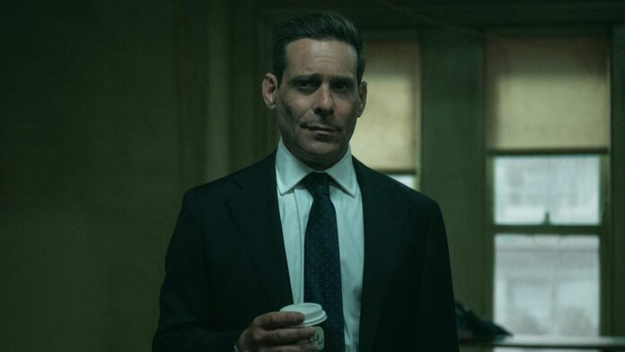 James Callis wears a suit and holds a cup of coffee while standing in front of two windows in Slow Horses season 5 episode 6