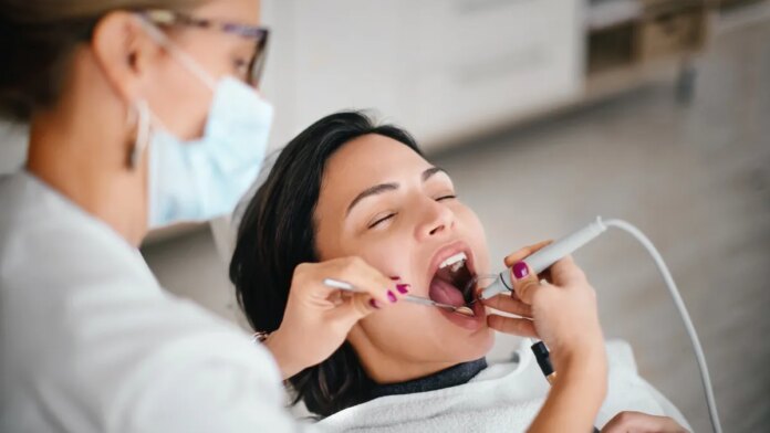 A dental hygienist with pulled-back blonde hair, glasses and a blue face mask removing calculus from a patient&apos;s teeth with a scaling tool. The patient has dark brown hair.