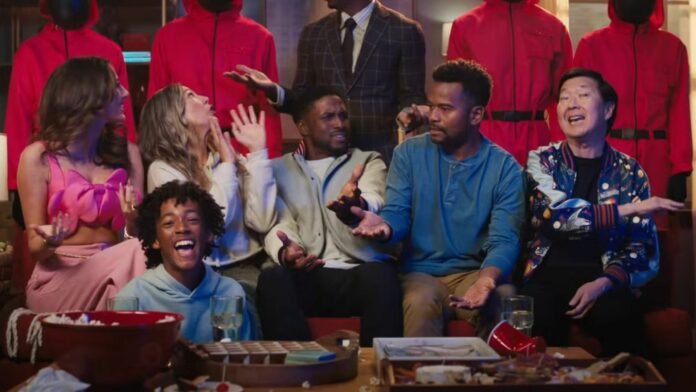 A group of people animatedly sit on a couch with Squid Game guards behind them